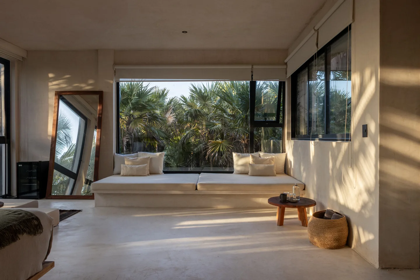 A room with a white bench, a mirror, and a view of palm trees.