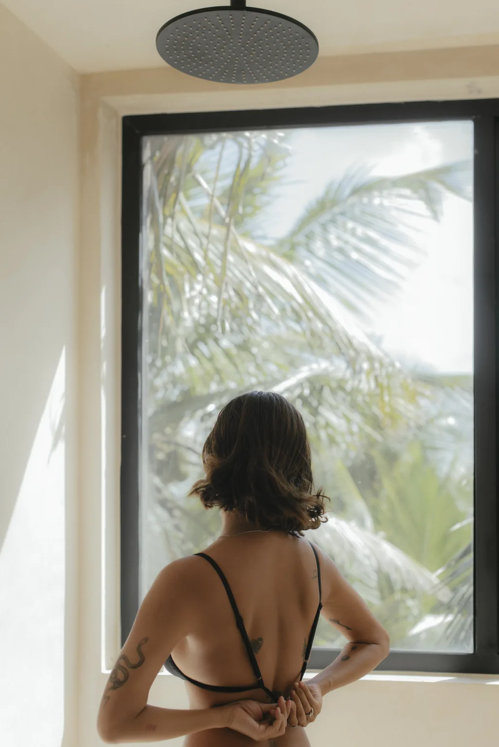 A woman with tattoos stands in front of a window in a bathroom.
