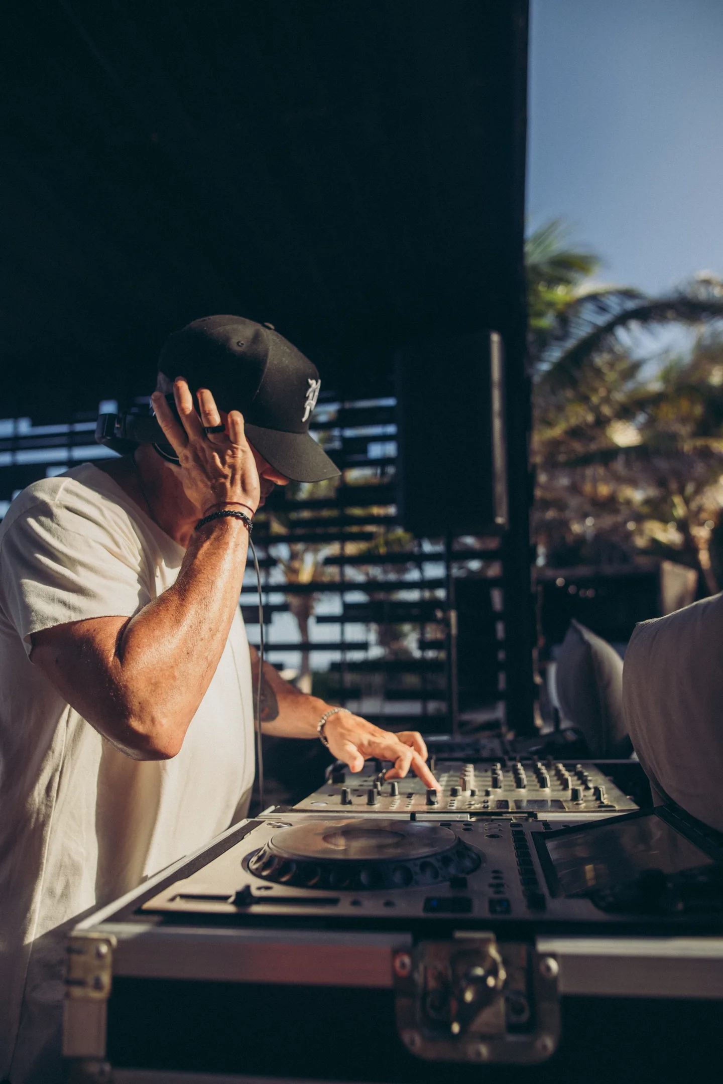 A man in a white shirt and black hat is DJing outdoors.